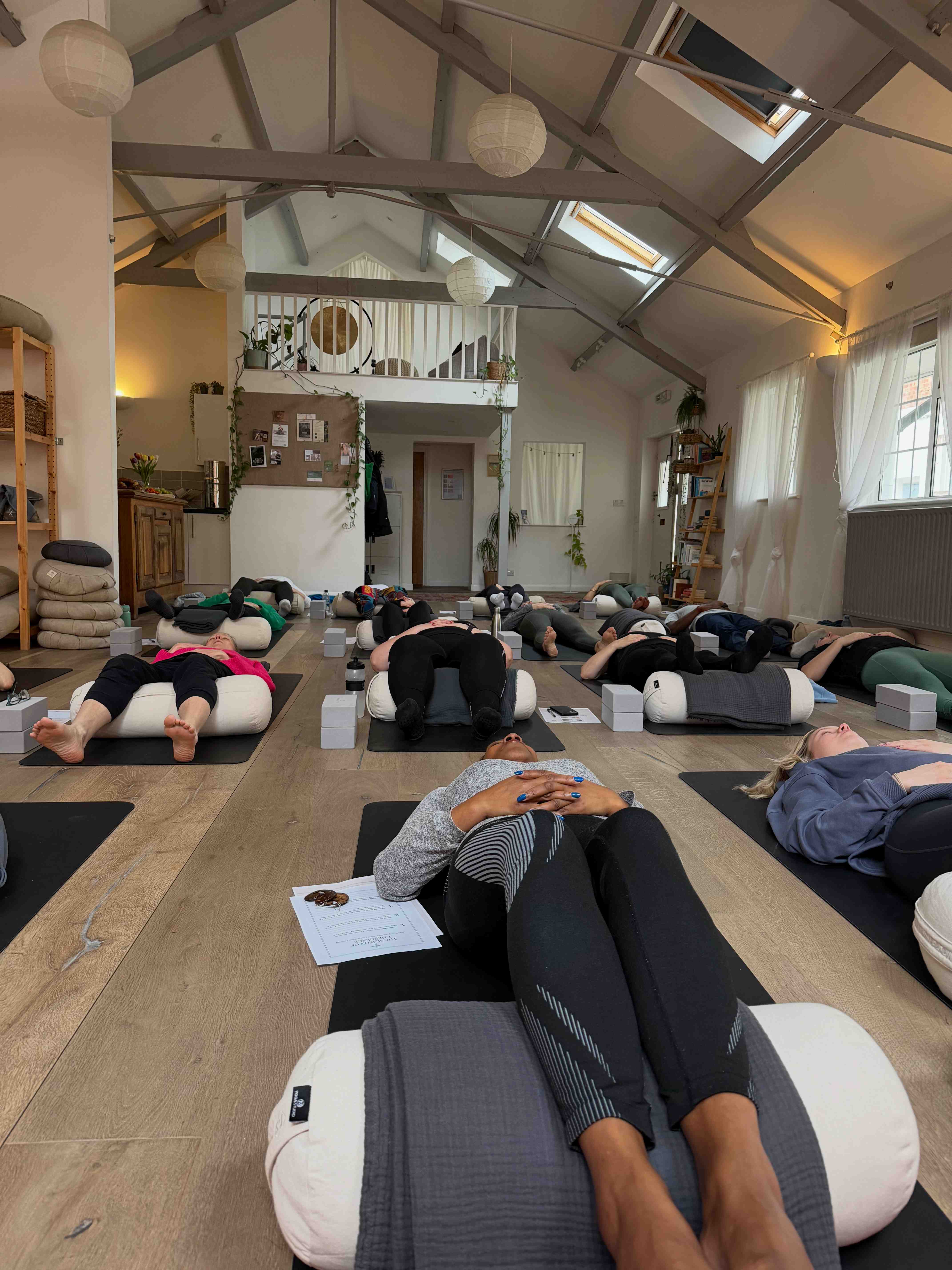 A group of people lying in a restorative yoga pose on bolsters in a warm, light-filled studio