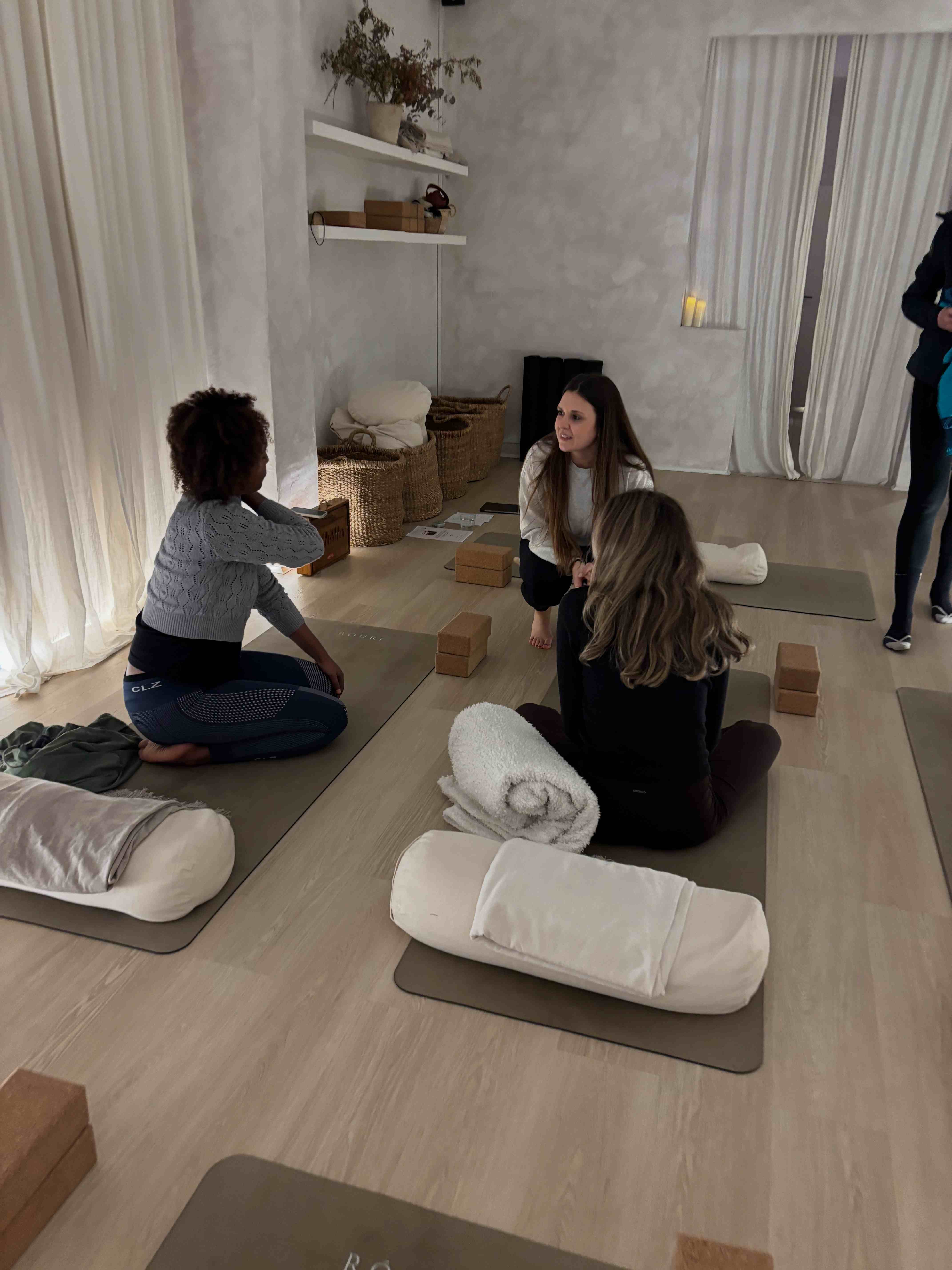 Three people seated in a small circle on yoga mats in a calm, white-draped studio